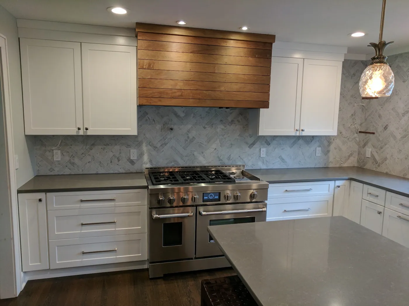 Professional gas range under a custom stained wood hood with marble herringbone backsplash and white shaker cabinets in a Monmouth County, NJ kitchen remodel